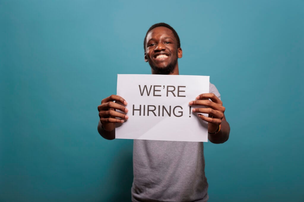 a young man smiling and holding up a printed paper written on it we're hiring